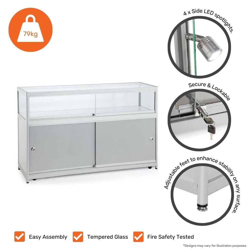 A silver aluminium glass counter display cabinet featuring a lockable storage compartment and four side LED spotlights. The top section is enclosed with tempered glass for secure product display. The cabinet includes adjustable feet for enhanced stability on any surface and a secure lockable door. The total weight of the unit is 79kg. Additional icons highlight its easy assembly, tempered glass construction, and fire safety testing. The design may vary slightly from the illustration.