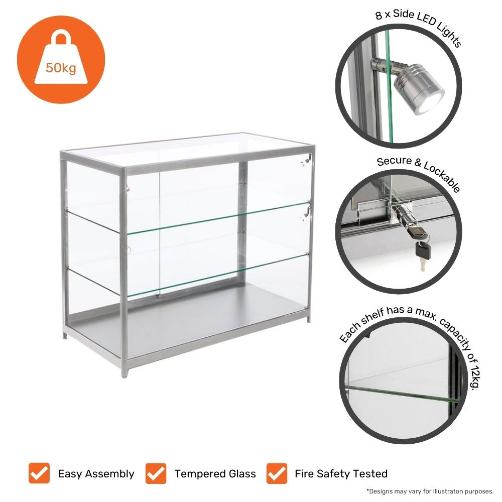 A silver glass counter display cabinet with a lockable sliding door, featuring tempered glass shelves and an aluminium frame. The cabinet is equipped with 8 side LED lights for enhanced visibility. Each glass shelf has a maximum weight capacity of 12kg, with a total load capacity of 50kg. The unit is designed for easy assembly and has been fire safety tested. Icons at the bottom highlight key features such as tempered glass, fire safety, and easy assembly.